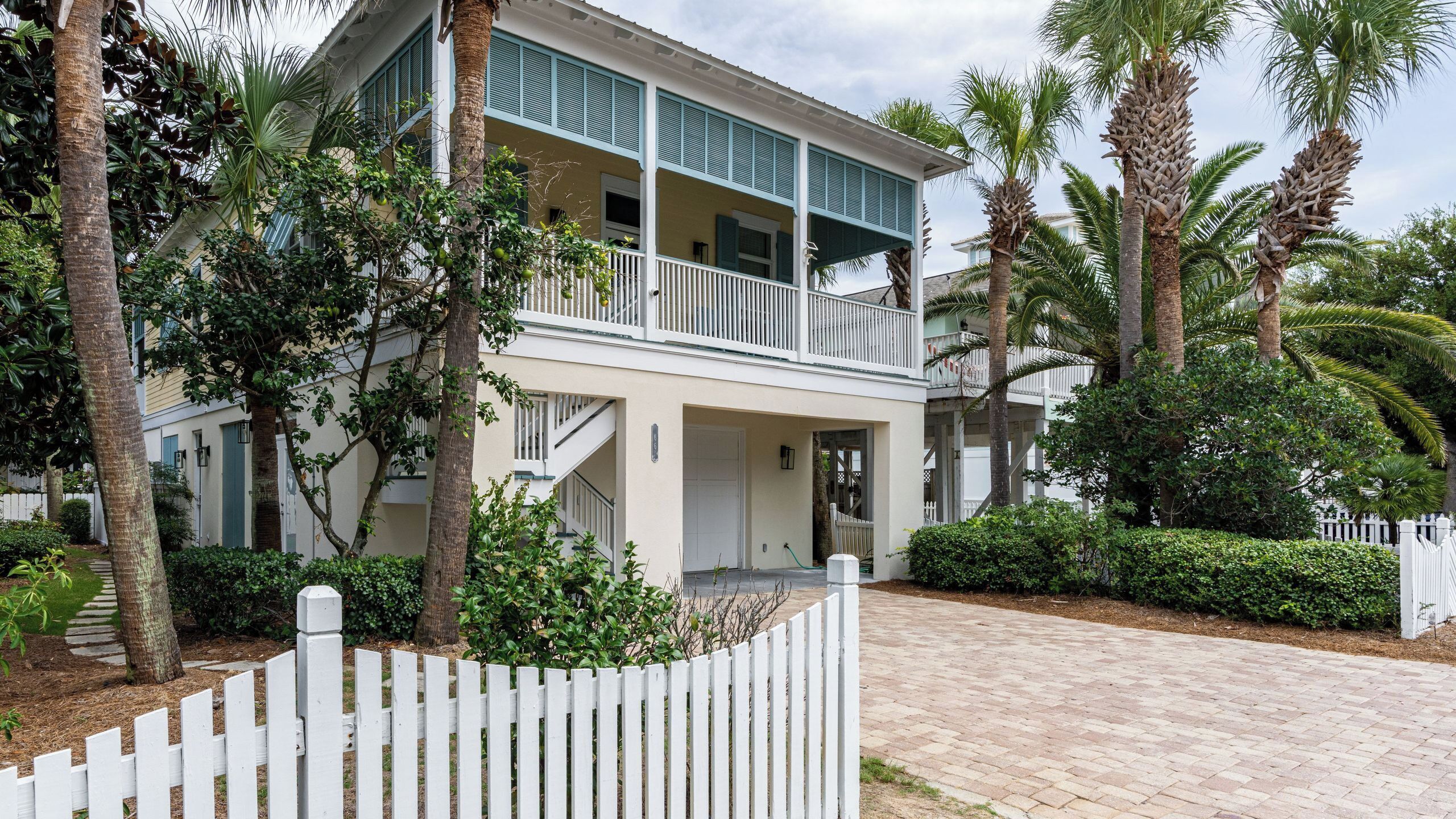 A two-story house with a balcony, surrounded by palm trees and a white picket fence.