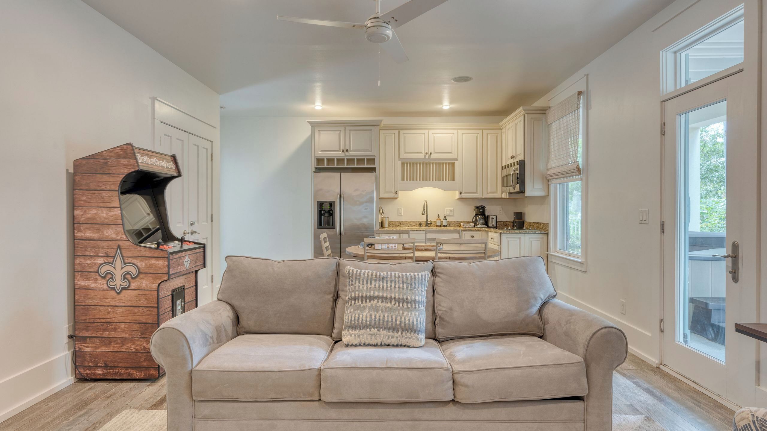Una sala de estar con un sofá color beige, un mueble de madera con un diseño de fleur-de-lis, y una cocina al fondo.