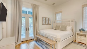 Un dormitorio con cama, un banco, una mesita de noche, televisor y ventana con cortinas.