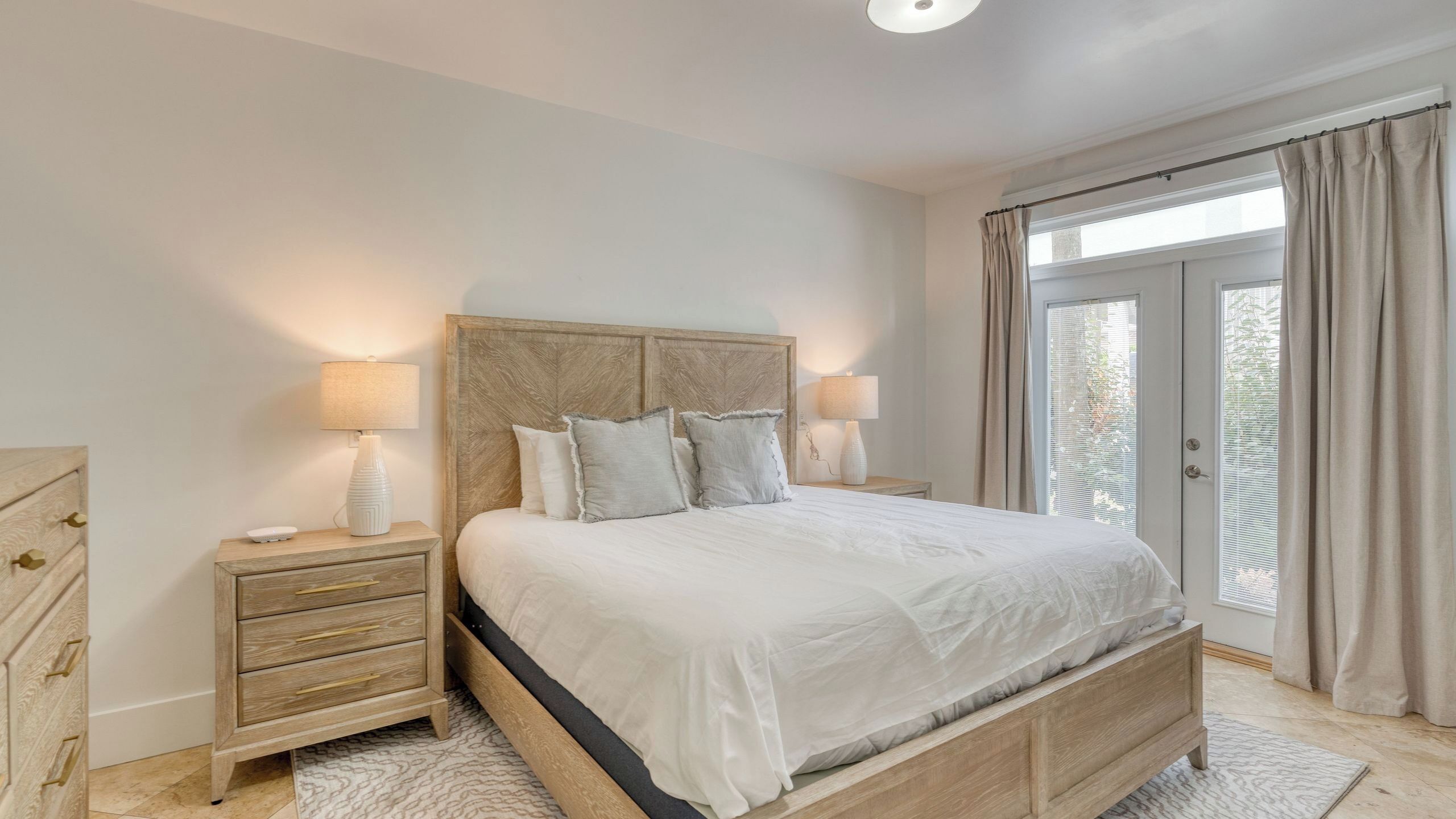 Une chambre à coucher avec un lit en bois, des tables de chevet, une commode, une fenêtre avec des rideaux et une vue sur la verdure à l’extérieur.
