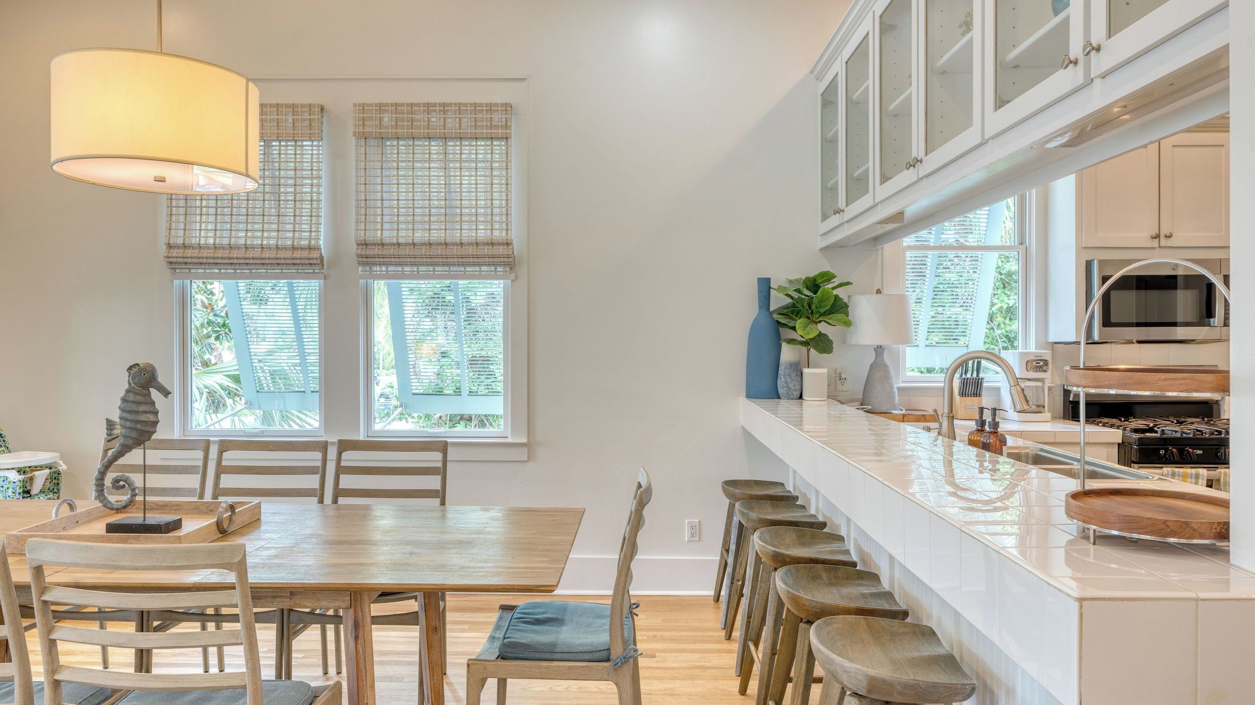 A modern kitchen with a central island, bar stools, a dining table, and large windows with blinds.