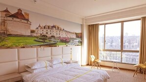 Une chambre d’hôtel avec un grand lit, un tableau représentant un château, une vue sur des immeubles par une grande fenêtre et une petite table avec des chaises.