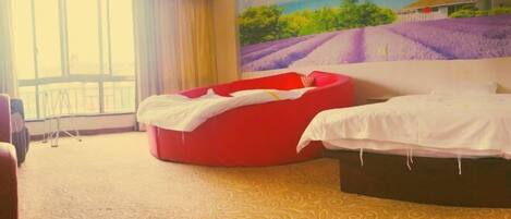 Una habitación con un ventanal grande, un sillón de peluche rojo, una cama y un mural con una casa en una colina.