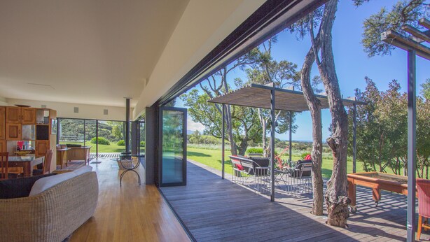 Un espace intérieur vaste et lumineux, doté d’un plancher en bois, d’un canapé et d’une table à manger. De grandes portes vitrées donnent accès à une terrasse extérieure où se trouvent une table de billard, des chaises et d’où l’on peut admirer un paysage verdoyant.
