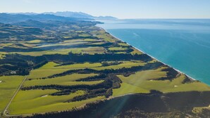 Vue aérienne d’un paysage côtier, avec des champs verdoyants vallonnés, une côte et des montagnes à l’horizon.