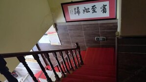 Un escalier recouvert d’une moquette rouge et doté de balustrades en bois, menant à une pièce dont un mur est recouvert de carreaux et sur laquelle est affiché un panneau dans une langue étrangère.