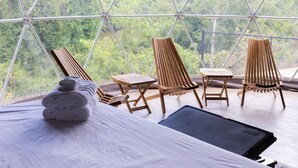 Una habitación con cama, una mesa de madera y tres sillas de madera. La habitación tiene ventanas amplias con vista a árboles verdes.