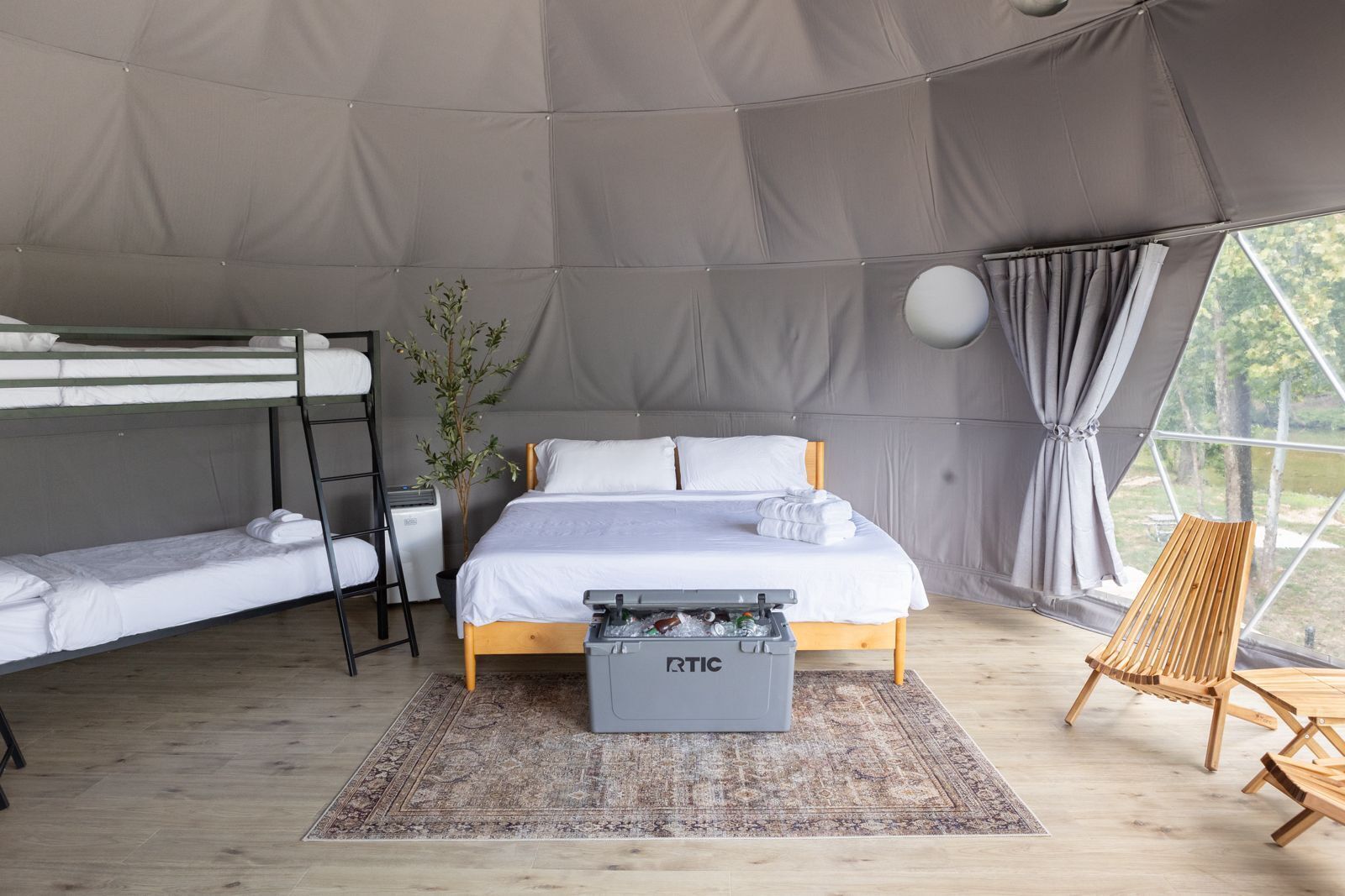 A tented accommodation room with bunk beds, a bedside table, a plant, a storage box, and a view of the outdoors.