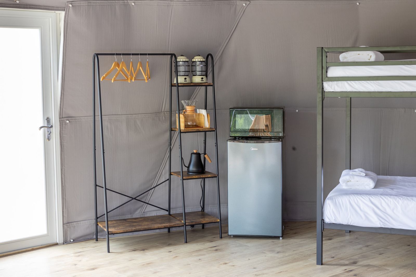 A compact room with a bunk bed, a kitchenette with a fridge and microwave, and a shelving unit with various items.
