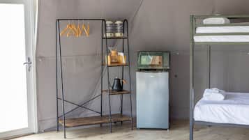A compact room with a bunk bed, a kitchenette with a fridge and microwave, and a shelving unit with various items.