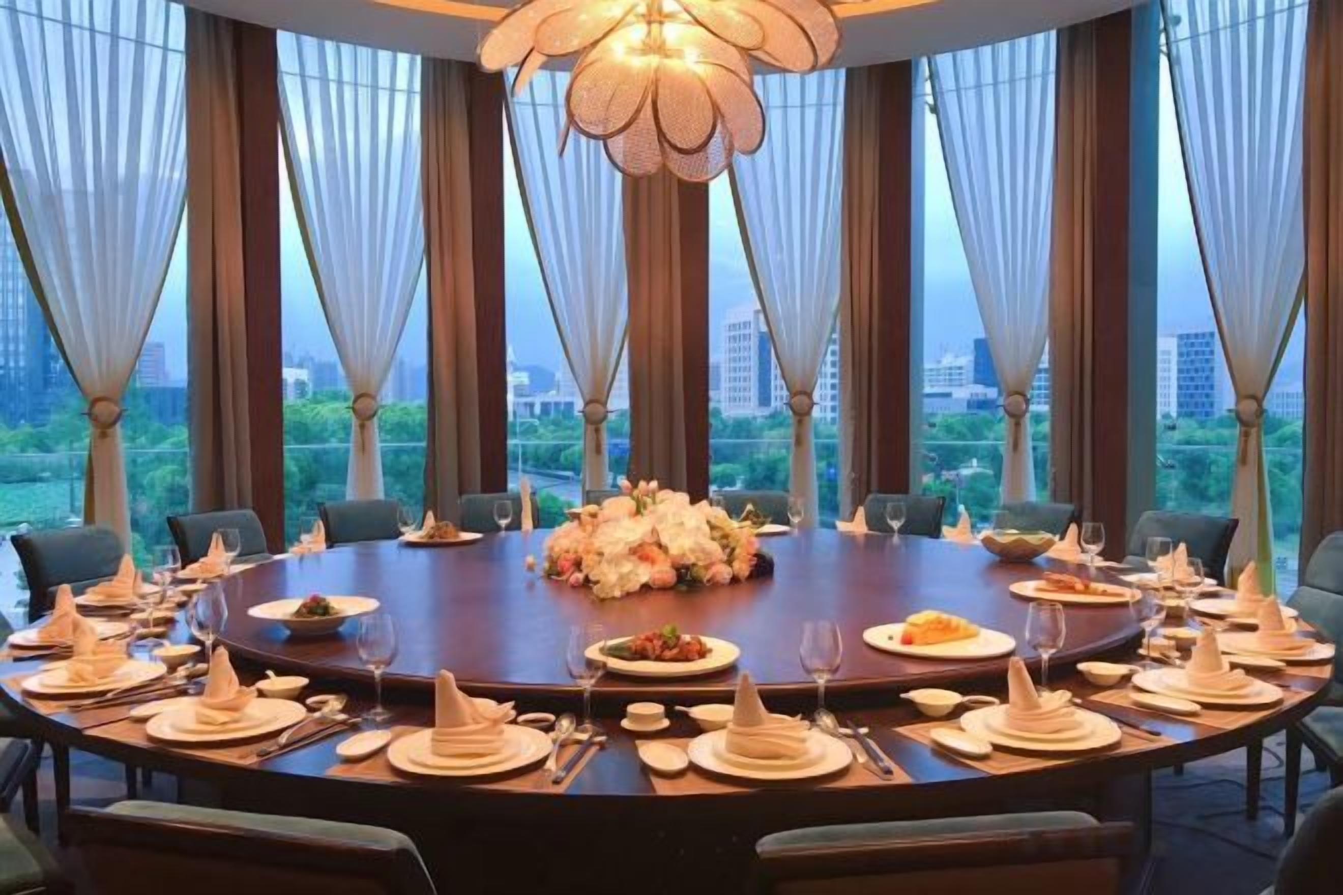 A round dining table set for a meal with a floral centerpiece, surrounded by chairs, in a room with large windows and a chandelier.