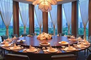 Una mesa de comedor redonda dispuesta para una comida, con un centro de mesa floral, rodeada de sillas, en una habitación con ventanas grandes y una araña.