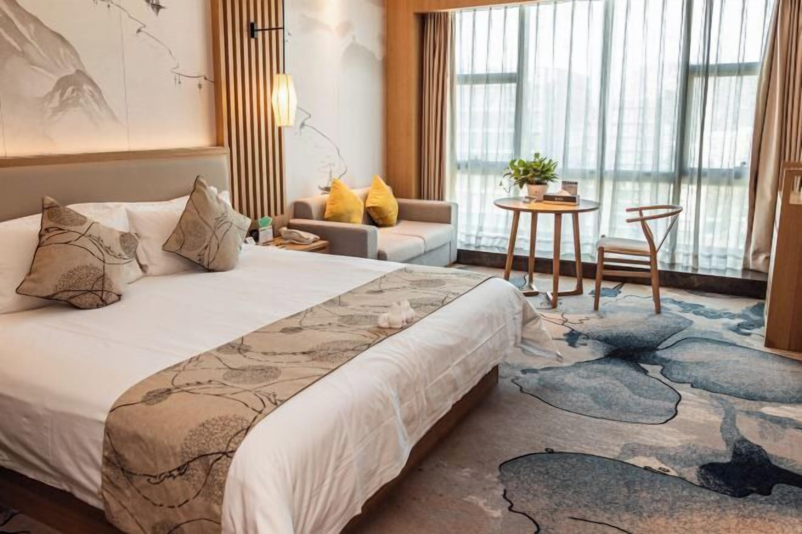 Habitación de hotel con cama, sofá, una pequeña mesa con sillas, una planta en maceta y vistas a través de amplias ventanas.