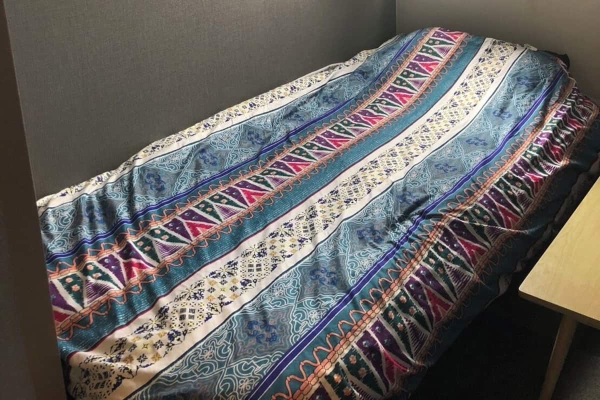 Una habitación pequeña con una cama individual, una colcha estampada, una ventana con cortinas y un estante con un gancho.