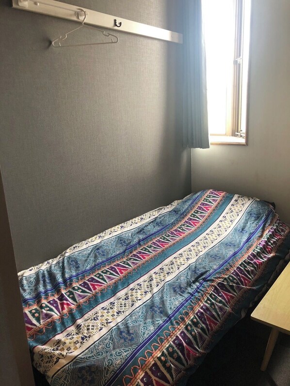 A small, single-bed room with a patterned bedspread, a window with curtains, and a wall-mounted shelf with a hook. - Guest house Blue Ocean Naha - Hostel (Naha)