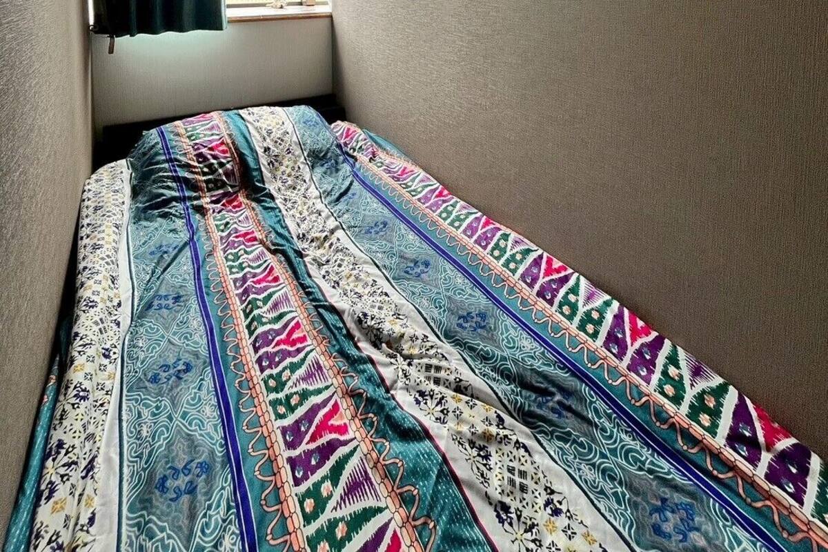 Una habitación pequeña y estrecha con una cama individual, una ventana con cortina y una lámpara fijada a la pared.