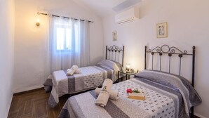 A hotel room with two beds, a nightstand, a window with curtains, and an air conditioning unit. - Casa Rueapara Torrox by Ruralidays (Málaga)