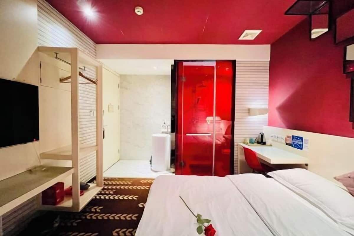 Habitación de hotel con cama, televisión, escritorio, zona de ducha y pared de acento roja.
