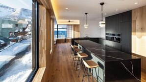 A modern kitchen with a black marble island, wooden bar stools, and a view of a snowy landscape through large windows. - Dharma Niseko (Kutchan)
