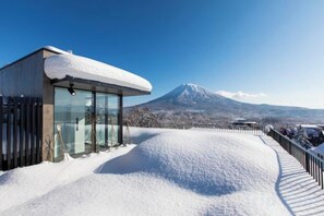 Schneebedeckte Landschaft mit einem modernen Gebäude, das große Fenster und eine Glaskonstruktion besitzt und auf einen Berg blickt.