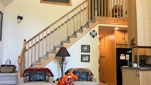 A living room with a staircase, a sofa, a coffee table with a vase, and a kitchen area with wooden cabinets and a refrigerator.