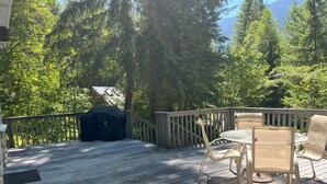 A wooden deck with a glass table and chairs, surrounded by trees and a mountain view.
