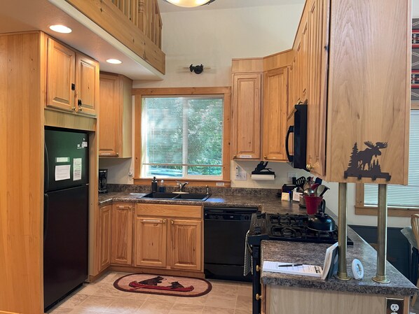 A kitchen with wooden cabinets, a black refrigerator, a stove, and a window with blinds. - Charming 2-bedroom cabin with AC and WiFi in wonderful Ashford (Ashford)