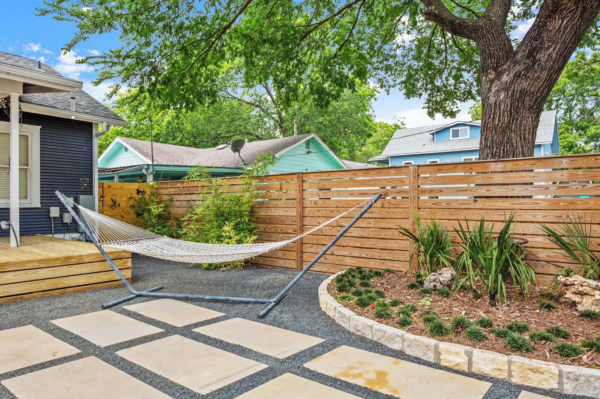 A hammock in a backyard with a wooden fence, a small garden, and a house in the background.