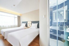 Quarto de hotel com duas camas de solteiro, box de vidro para o chuveiro e uma grande janela que proporciona iluminação natural.
