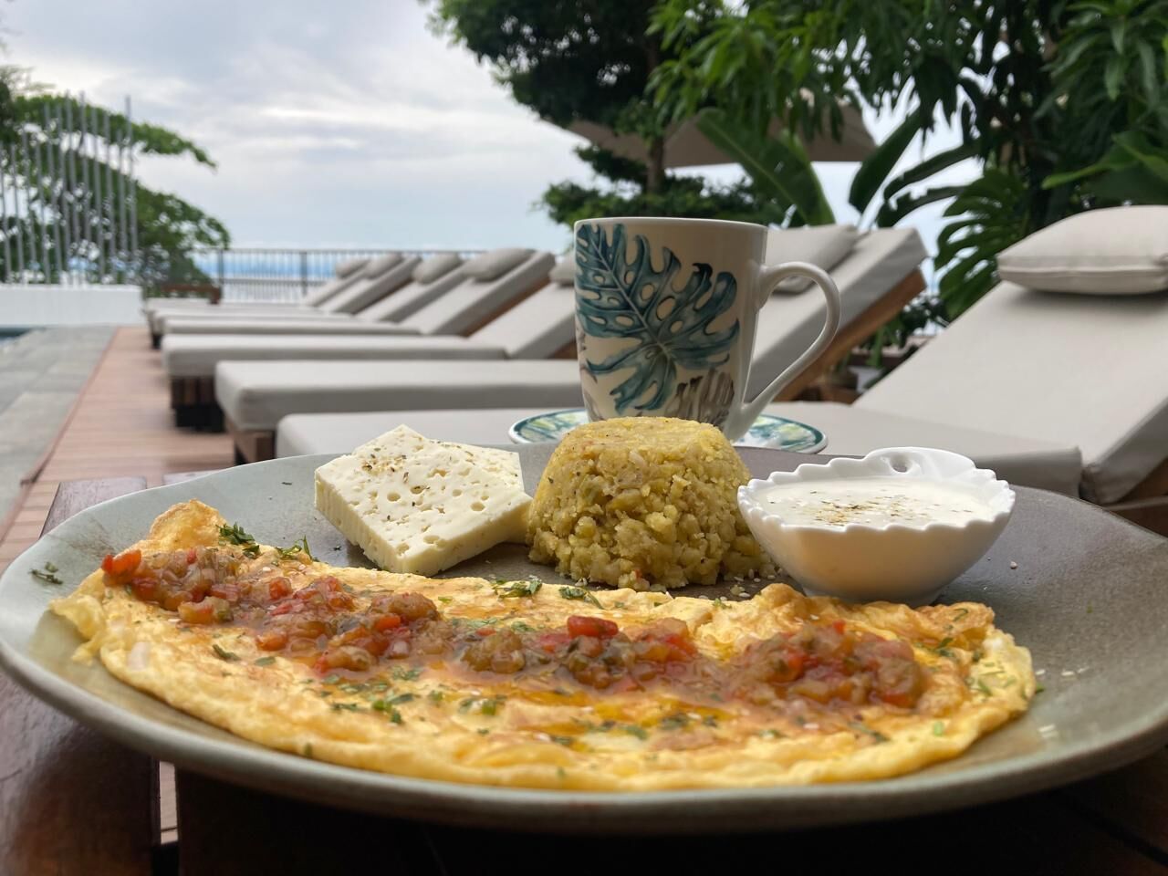 A plate with an omelette, a mound of rice, and two bowls of sauce on a table with a view of a pool and lounge chairs.