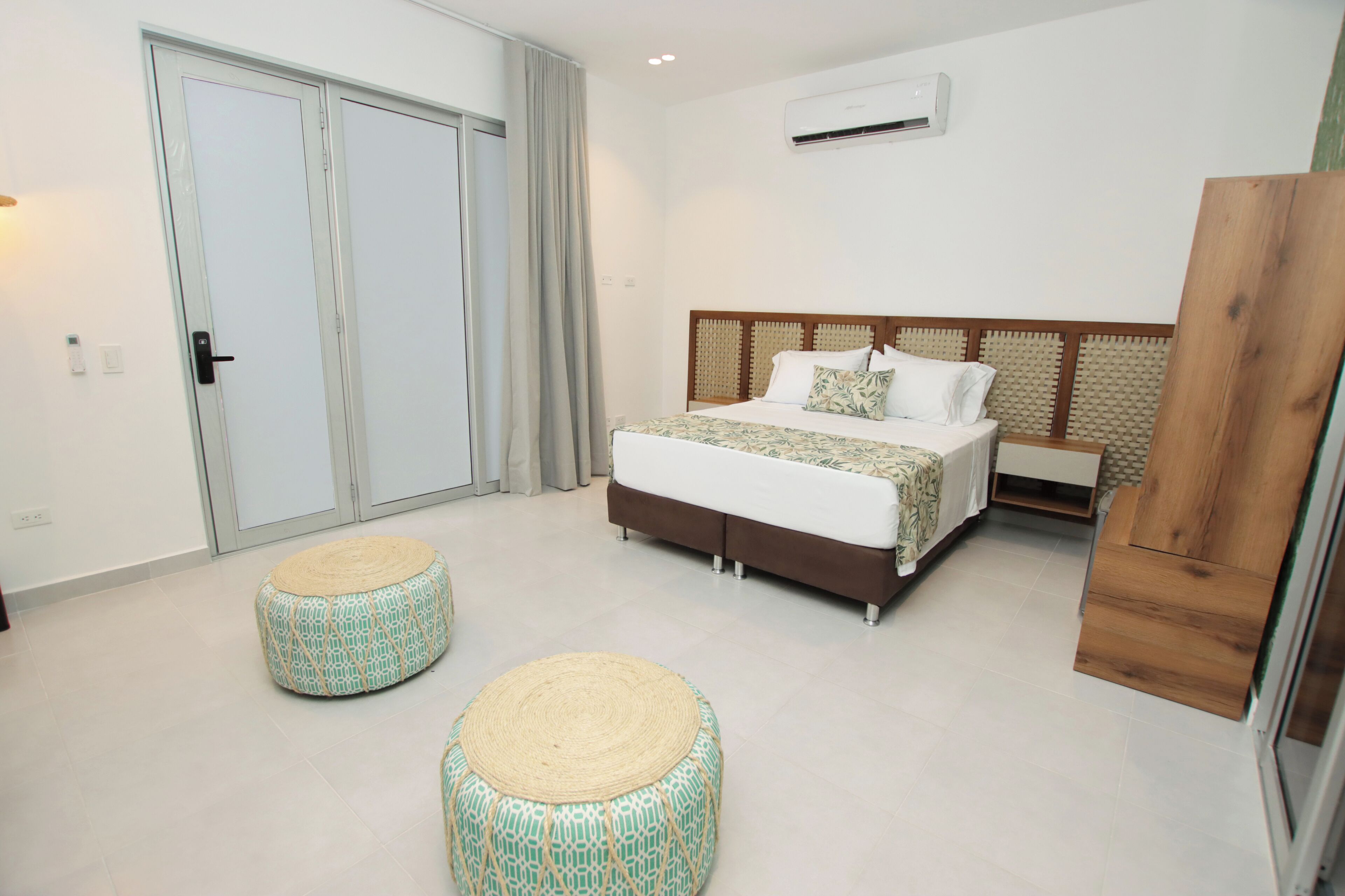 A bedroom with a bed, two ottomans, a wardrobe, and an air conditioning unit.