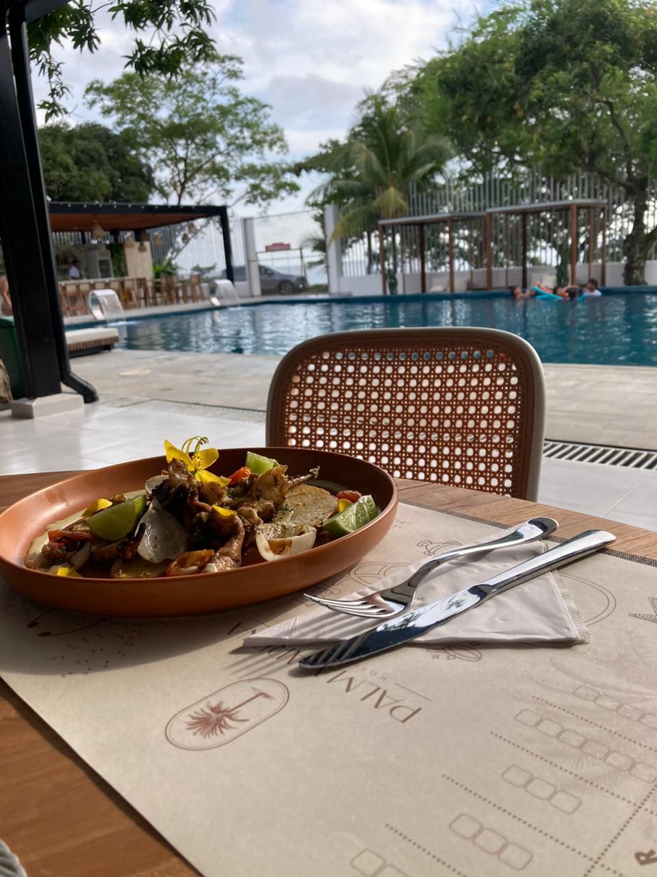Une table à manger avec une assiette de nourriture, une chaise et des couverts. Au fond, on aperçoit une piscine où des personnes nagent.
