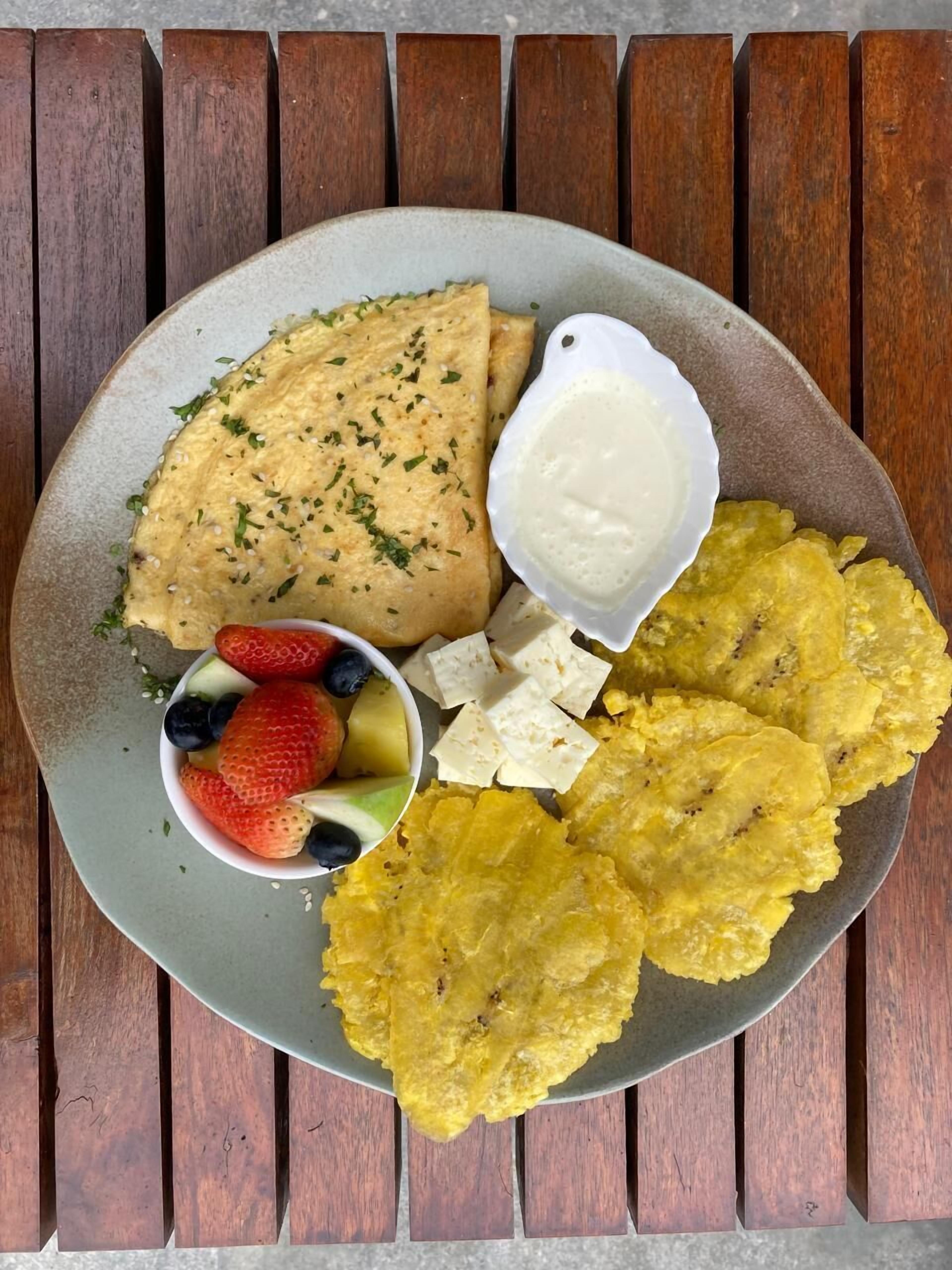 Un plato con una rebanada de tostada de aguacate, un bol de fruta, un recipiente pequeño de salsa blanca y plátanos fritos.