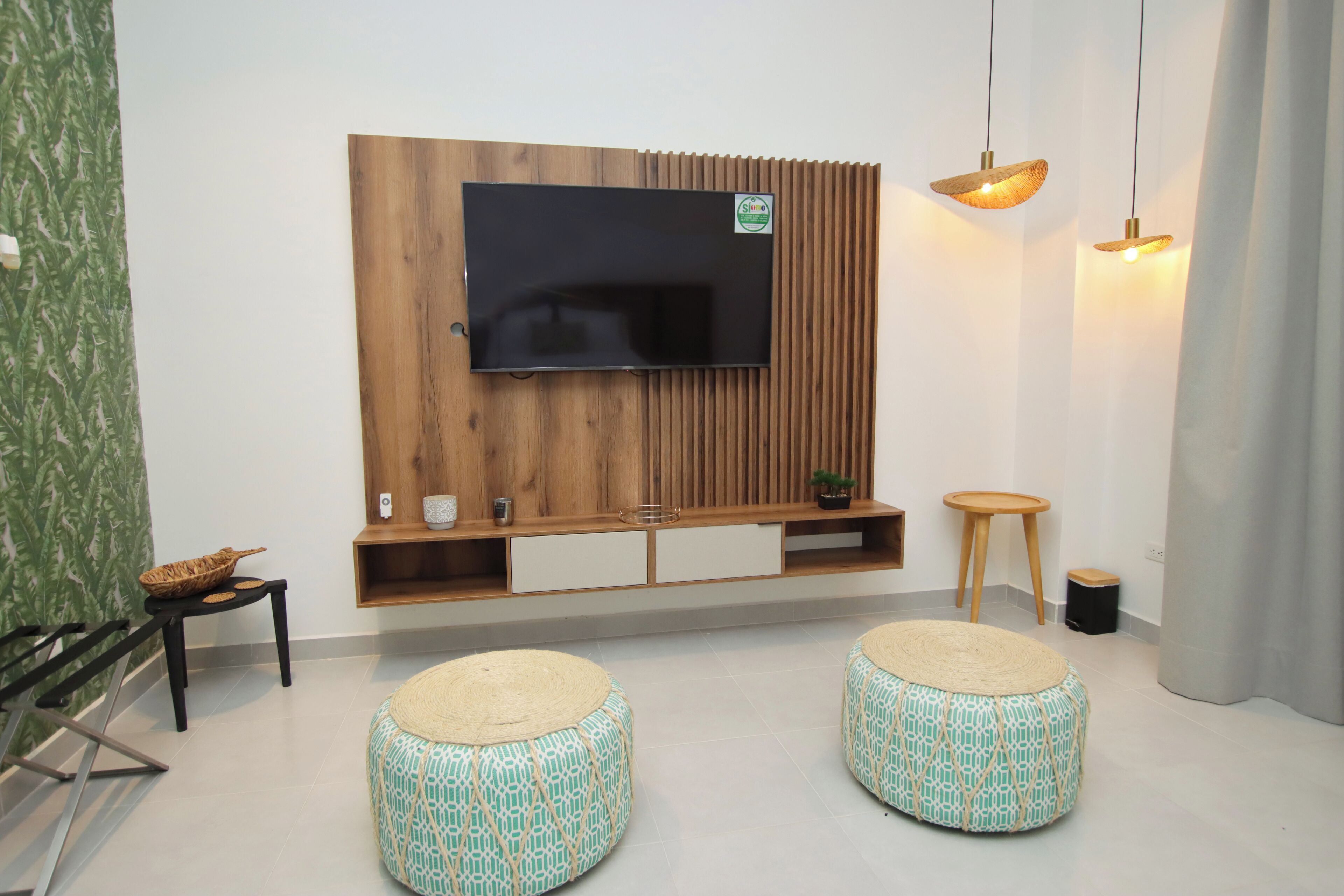 A modern living room with a flat-screen TV mounted on a wooden panel wall, two round ottomans with patterned cushions, a small wooden side table, and a potted plant.