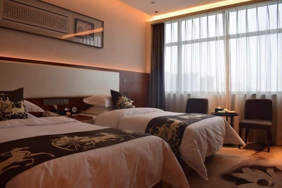 A hotel room with two beds, a large window with curtains, a small table, and a chair.