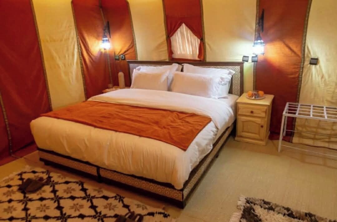 A bed with a white and orange bedspread, a bedside table with a lamp, and a small shelf with fruit.