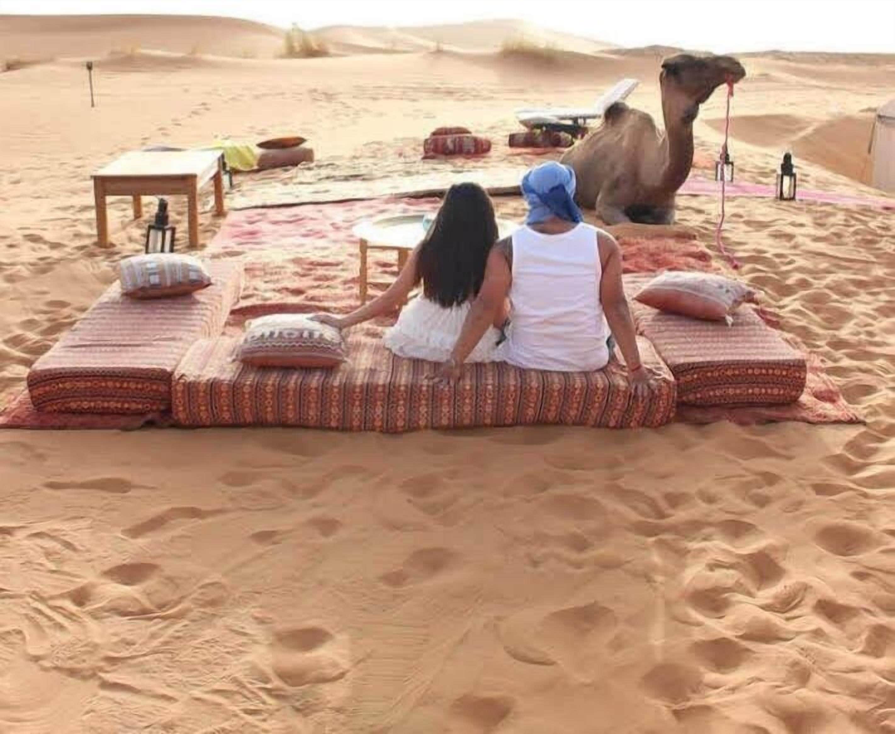Two people sitting on patterned cushions in a desert setting with a camel nearby.