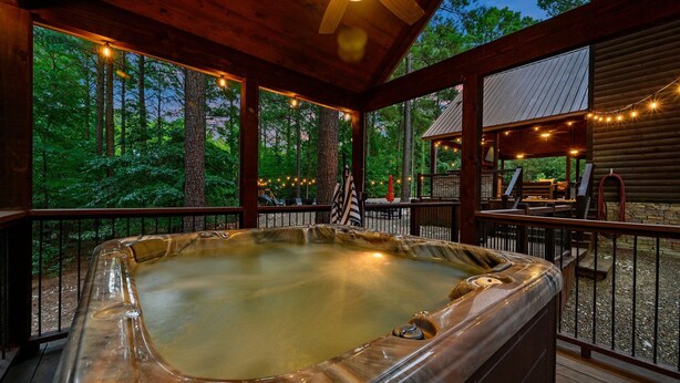 A hot tub on a wooden deck with string lights, surrounded by a wooden cabin and a forest view.