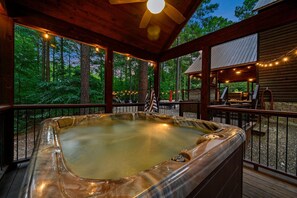 Een hottub op een houten terras, verlicht met lichtslingers, omgeven door een houten hutje en uitzicht op een bos.