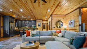 A spacious living room with a large L-shaped gray sofa, a round wooden coffee table, and a kitchen area with modern appliances and wooden walls.