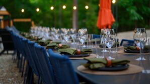 A dining table set with multiple wine glasses and plates, surrounded by blue chairs, in an outdoor setting with string lights in the background.