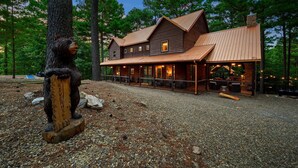 A rustic cabin with a large porch, a bear sculpture, and a gravel driveway.