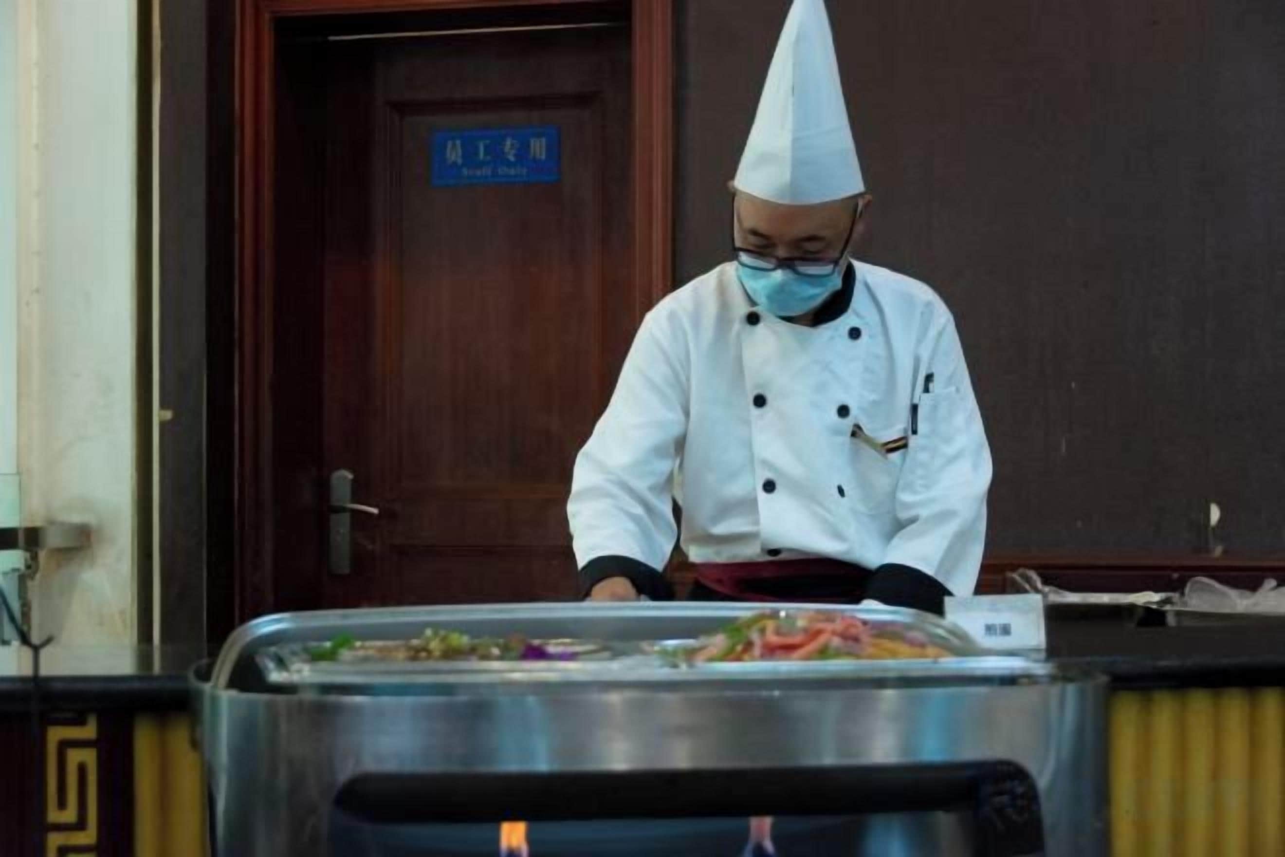 Un chef con uniforme y gorro blanco preparando comida en una cocina con mostrador de acero inoxidable y una puerta al fondo.