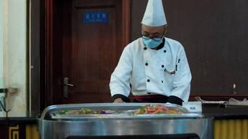 Un chef con uniforme y gorro blanco preparando comida en una cocina con mostrador de acero inoxidable y una puerta al fondo.