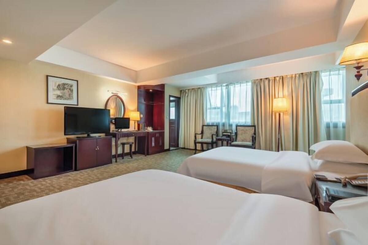 A hotel room with two beds, a desk with a chair, a television, and a large window with curtains.