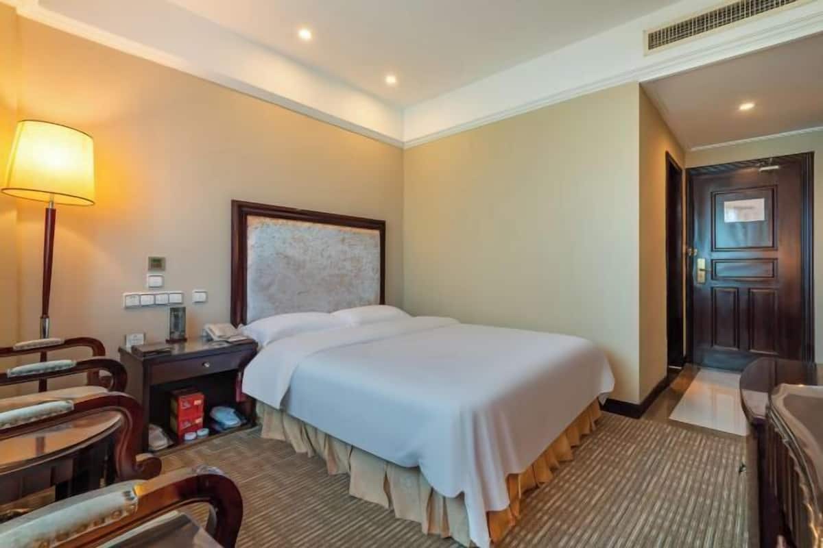A hotel room with a large bed, a desk with a lamp, a chair, and a small table.