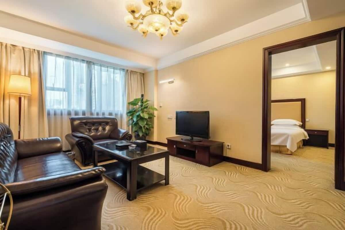 A hotel room with a leather sofa, a TV, a coffee table, a plant, and a bed with a white duvet.