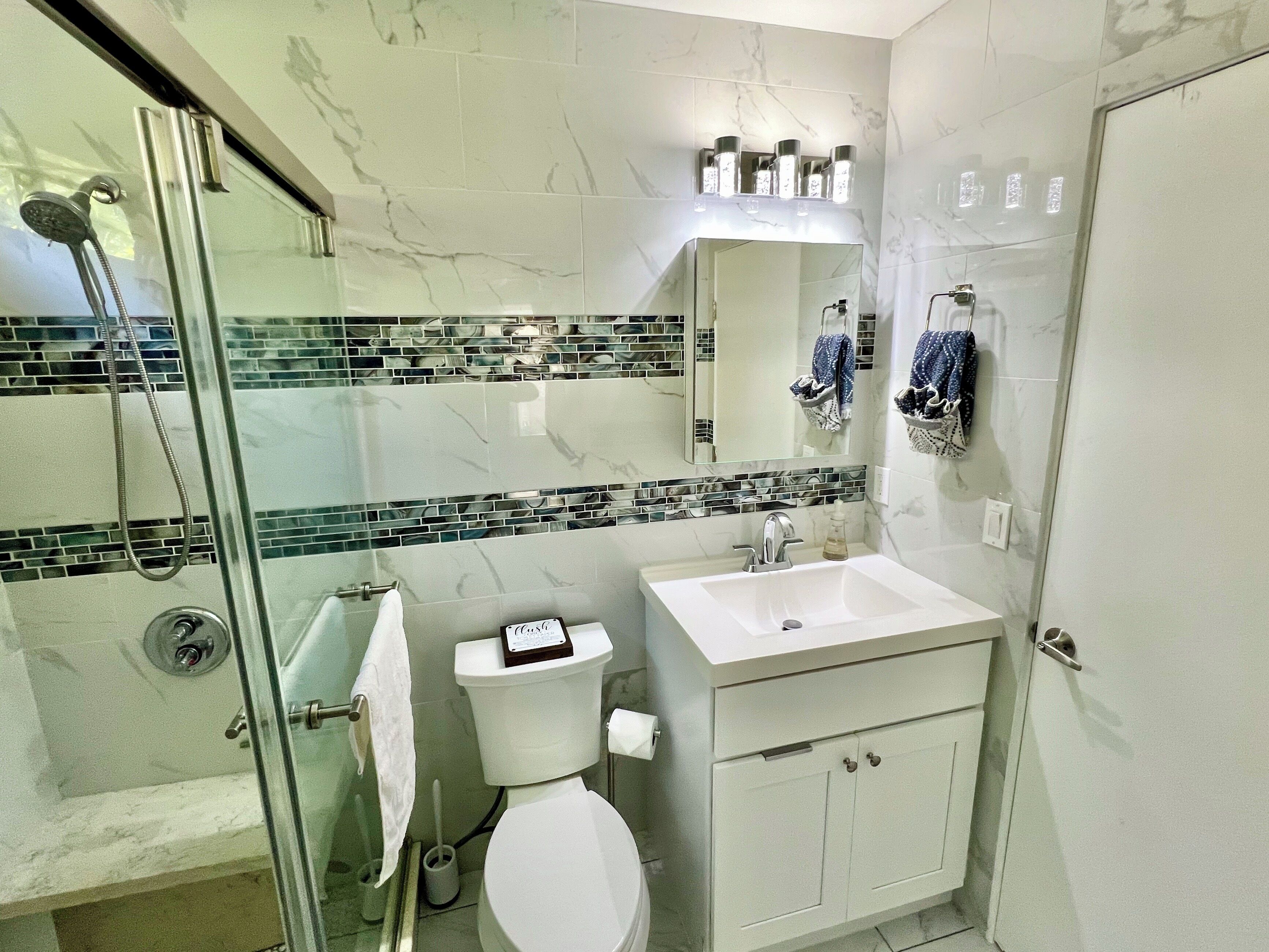 A bathroom with a glass shower enclosure, a white toilet, a white sink with a mirror above it, and a tiled backsplash.