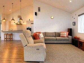 En moderne stue med sofa, lænestol og sofabord. I baggrunden er der et køkkenområde med spisebord og stole.