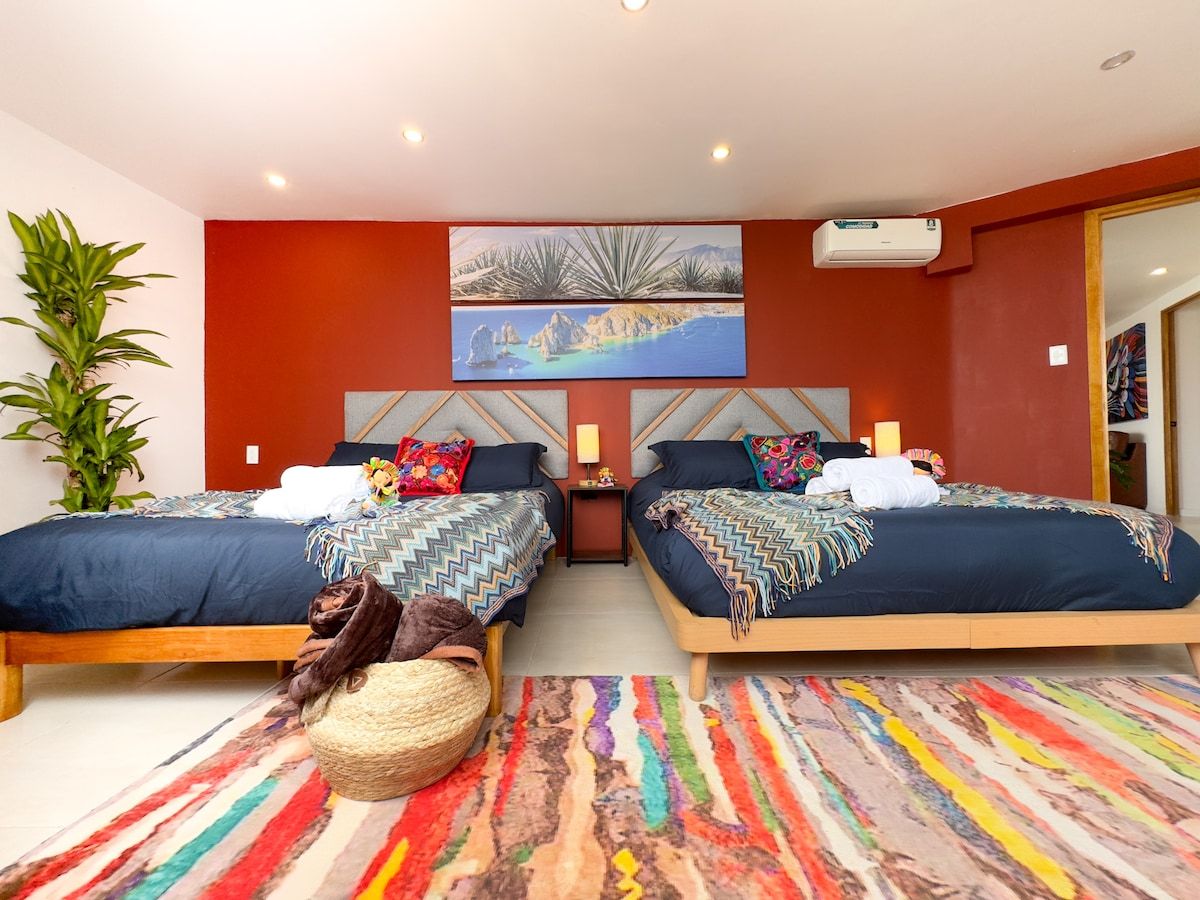 Une chambre double avec deux lits, un tapis coloré, un grand tableau et une plante en pot.